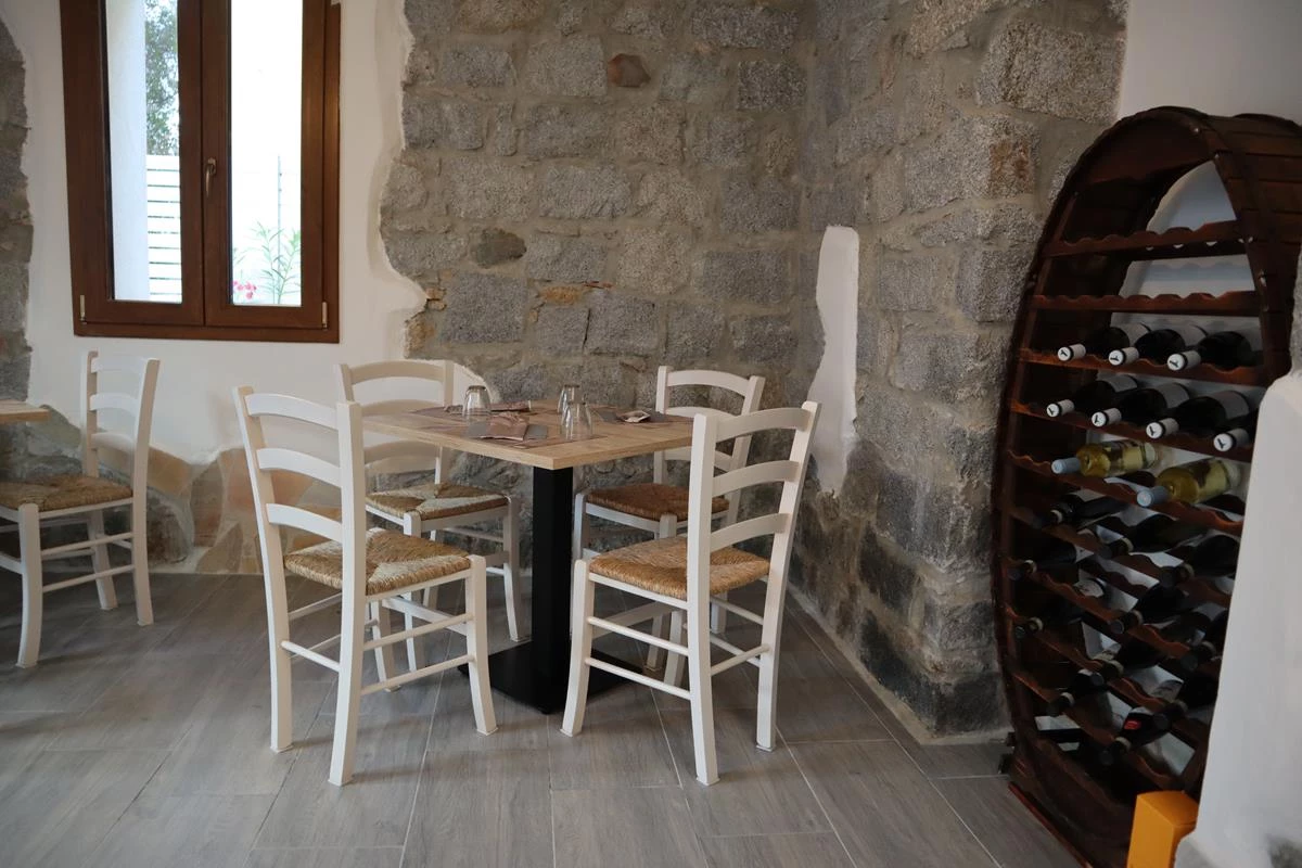 Sala di Zinnibiri con muro in granito, tavolo apparecchiato, sedie bianche impagliate e cantinetta a botte con vini