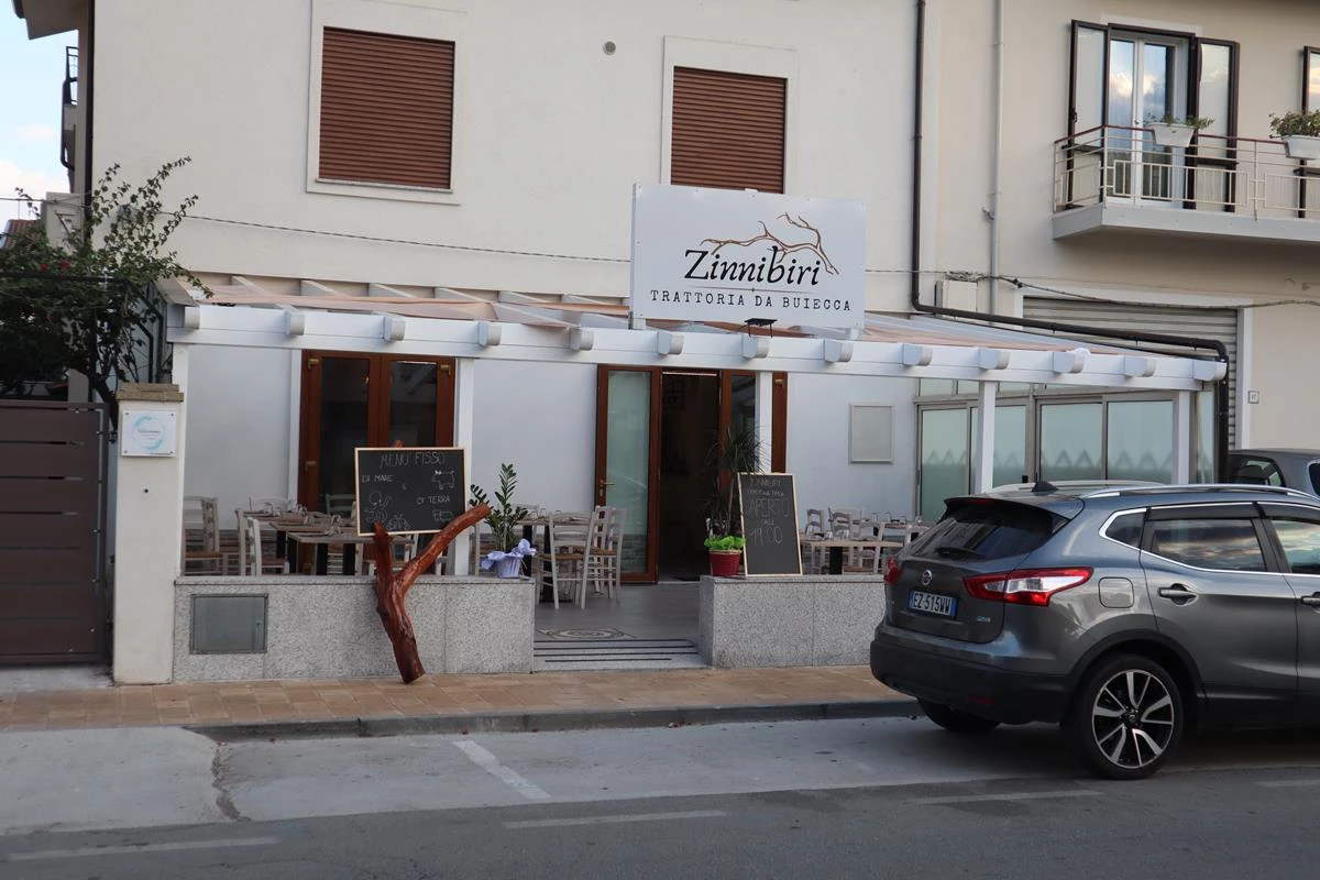 Ingresso con insegna «Zinnibiri Trattoria da Buiecca», veranda bianca, tavoli in legno e lavagne del menù fisso
